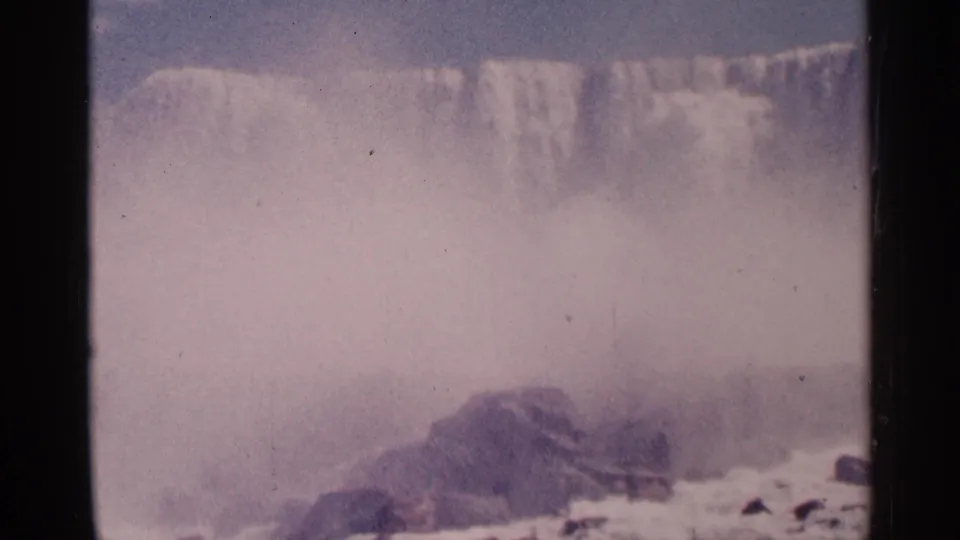 1964 Niagara Falls home-movie frame showing a broad cascade, dense mist, foreground rocks, and foaming water in faded color.