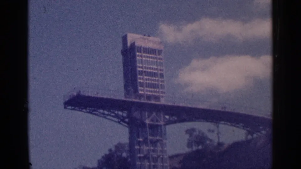 1964 8mm view near Niagara Falls showing a tall man-made observation or bridge-like structure, blue sky, clouds, and trees.