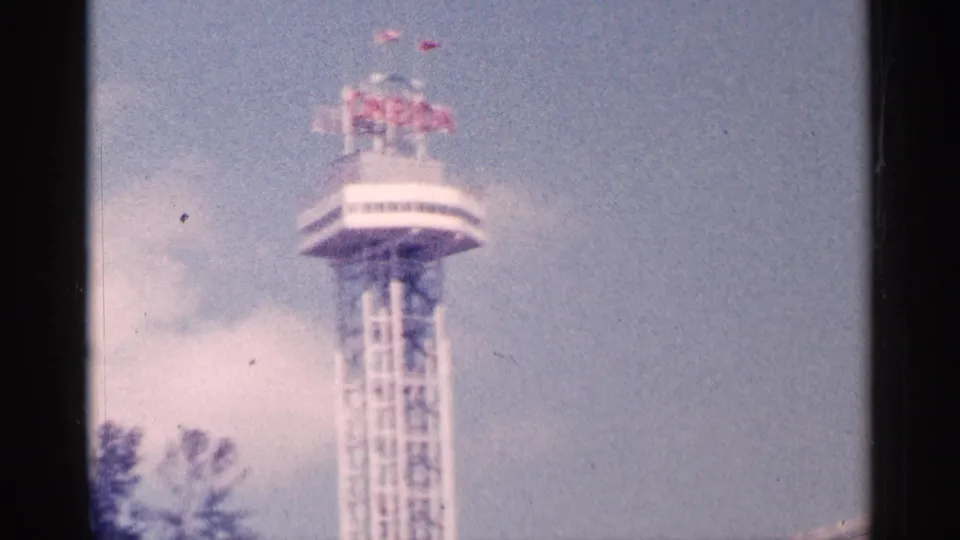 1964 Niagara Falls home-movie frame showing a tower with red rooftop lettering, a small flag, and an observation deck.