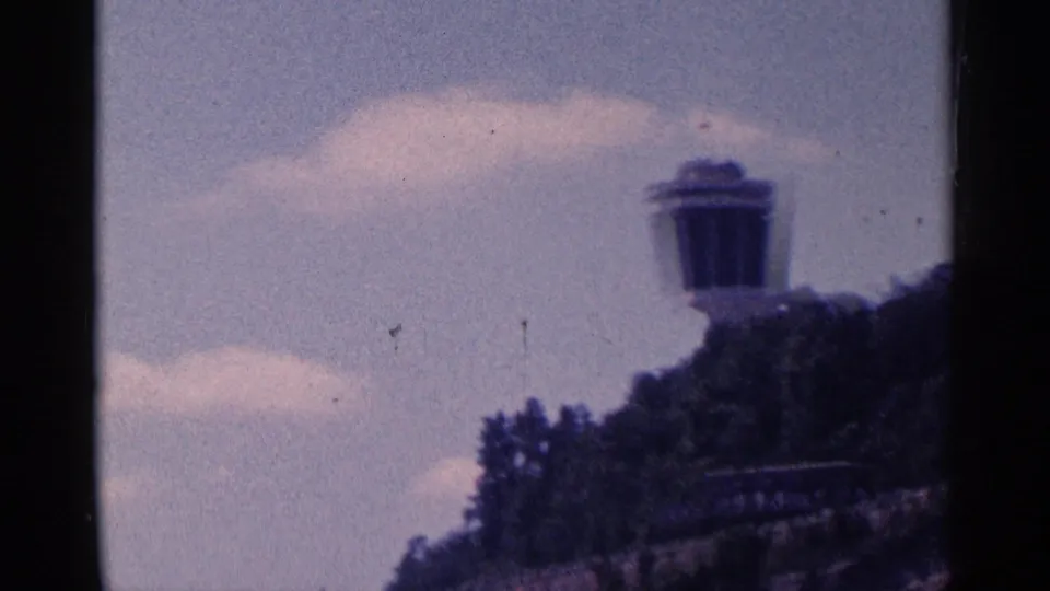 Wider 1964 home-movie view of the tower top above treetops, with hillside and clouds in the foreground and background.