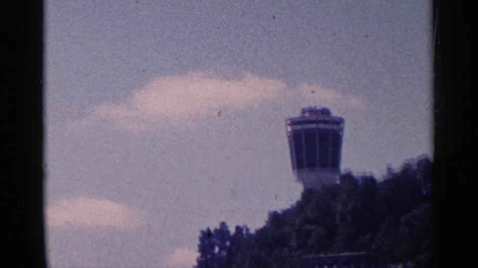 Final 8mm frame holds on the distant tower top above a dark tree line, with pale clouds behind it.