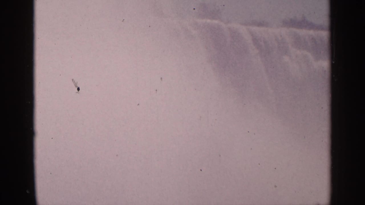 1964 8mm view of Niagara Falls in Canada, showing the waterfall edge through haze, with dust specks and dark film borders.