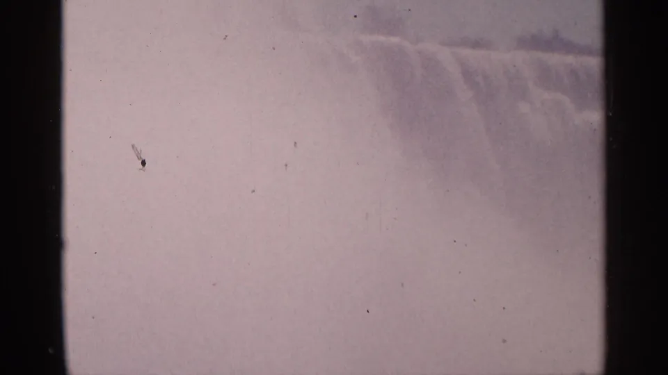 1964 8mm view of Niagara Falls in Canada, showing the waterfall edge through haze, with dust specks and dark film borders.