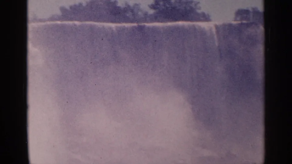 1964 8mm Niagara Falls view with broad falling water, mist, and a dark tree line in soft purple-tinted color.