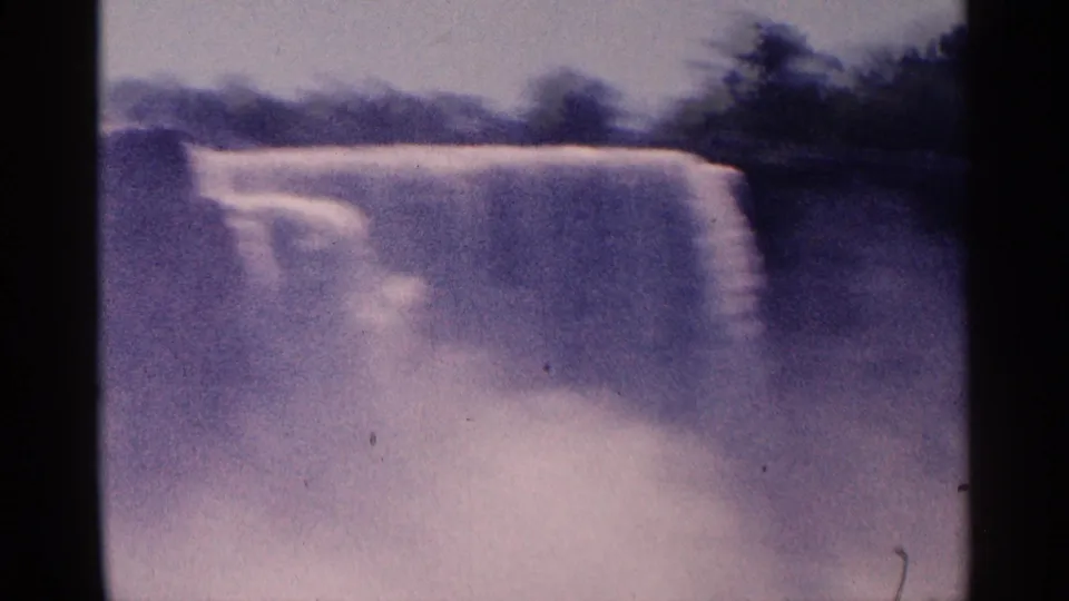 Soft, purple-tinted 8mm frame of Niagara Falls with a waterfall ledge, mist, and dark foliage beyond.