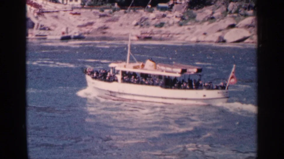1964 Niagara Falls tour boat carrying distant tourists across turbulent water below a rocky shoreline.