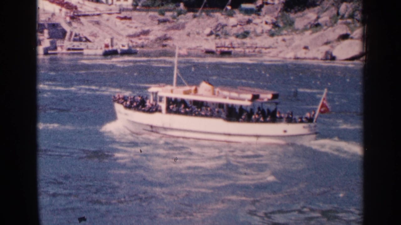 Crowded white tour boat moves across rough river water near a rocky shoreline.