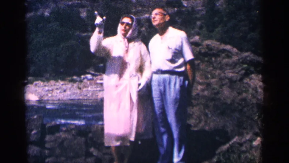 At Niagara Falls, Canada, the woman in a pink raincoat lifts one hand while the man stands beside her in a pale shirt and trousers.