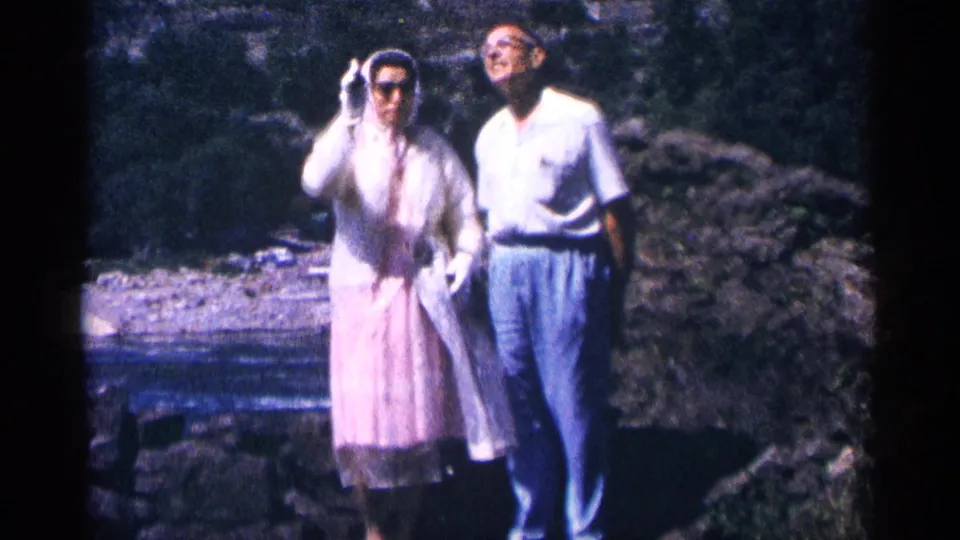 Mid-1960s home-movie frame showing the couple by the river as the woman adjusts her sunglasses or hood near Niagara Falls, Canada.