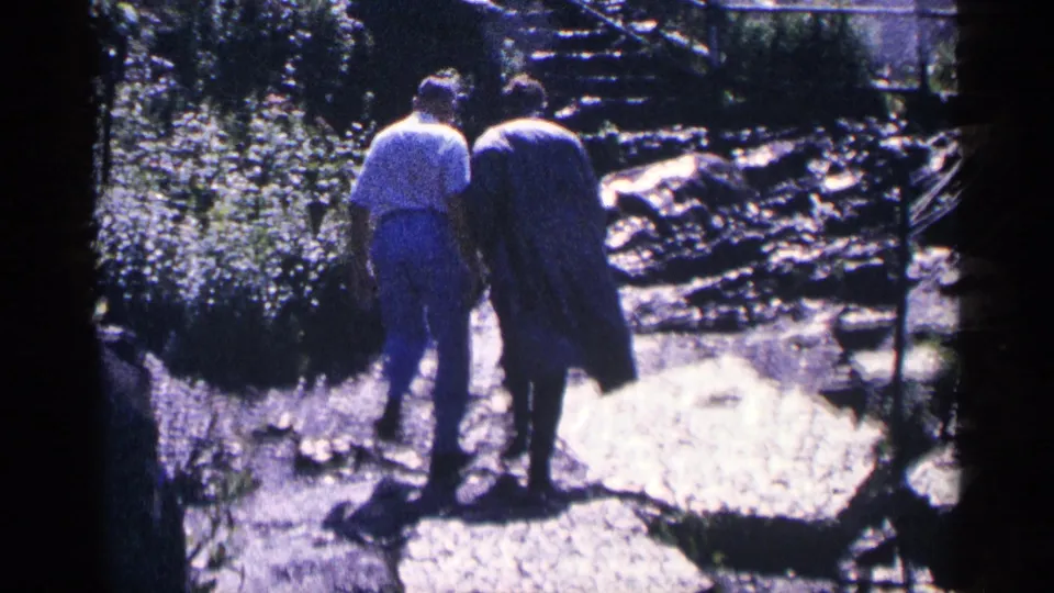 1964 home-movie frame near Niagara Falls, Canada, showing the couple walking away on a stone path through a landscaped riverside area.