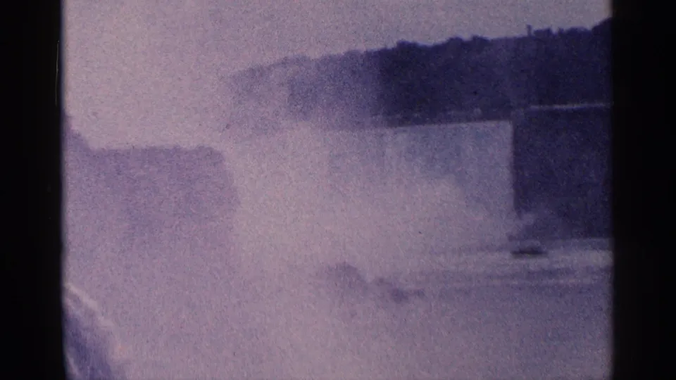 Vintage 8mm frame of Niagara Falls with dense spray, the river below, and a dark wooded ridge across the background.