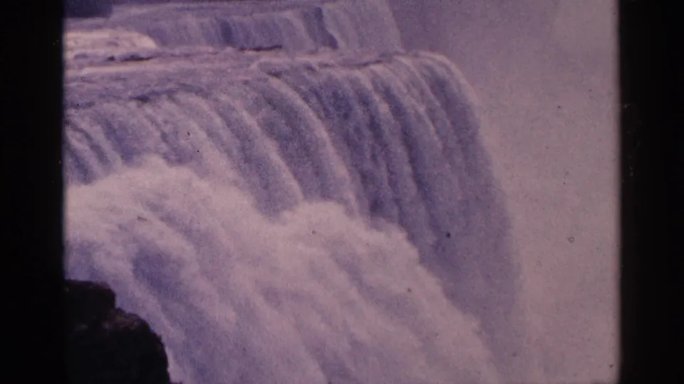 1964 8mm home-movie frame showing water cascading over Niagara Falls, with white spray and dark foreground rock at the edge.
