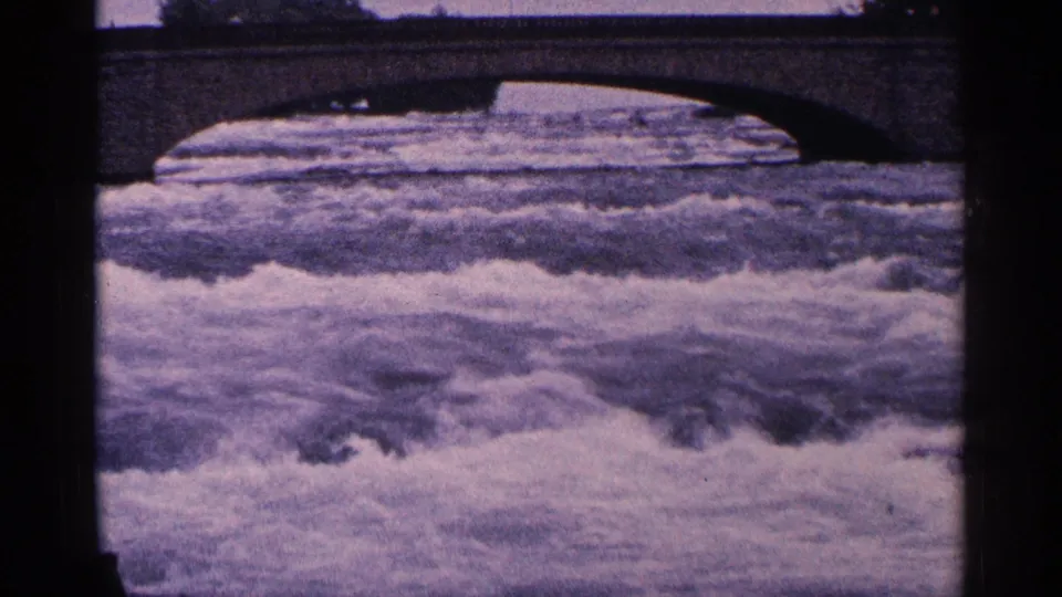 1964 8mm view of rough Niagara river water passing below a stone arch bridge, with heavy grain and dark frame edges.
