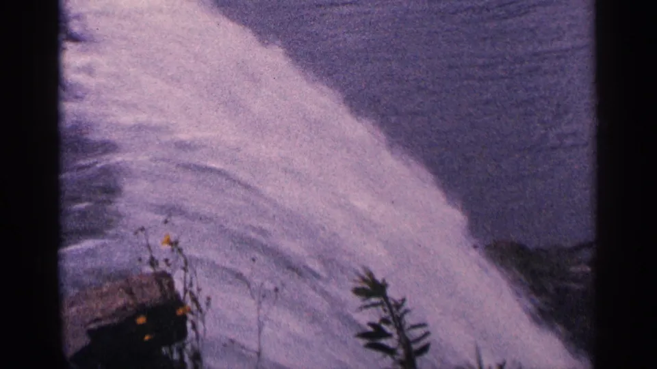 1964 Niagara Falls home-movie frame with a bright rushing water plume, dark river surface, and foreground vegetation.