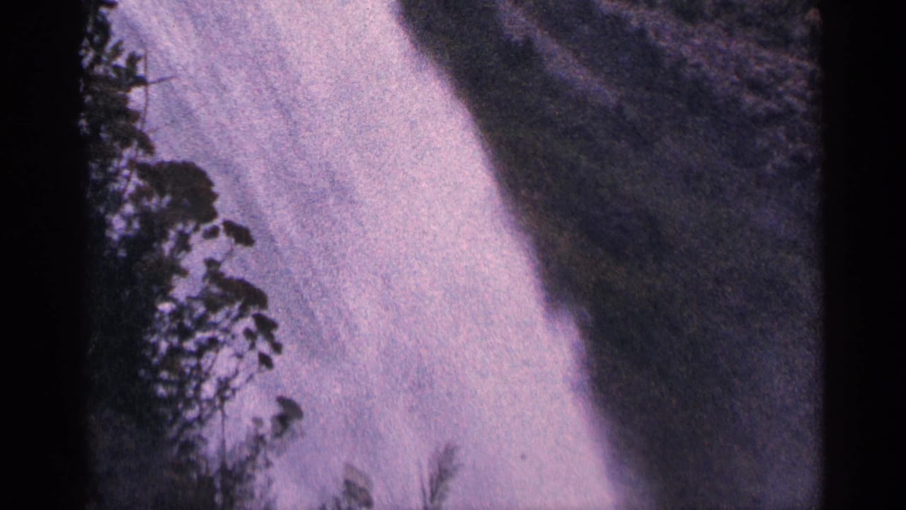 Vintage 8mm frame of Niagara Falls with pale rushing water, mist, and dark tree shapes near the center-left.