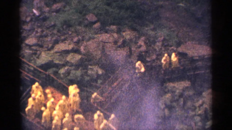 Distant workers or visitors in yellow outerwear stand along a rocky Niagara Falls riverbank in 1964 home-movie footage.