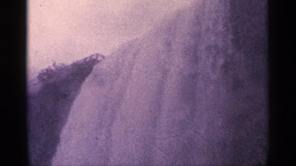 8mm frame from Niagara Falls, Canada, where pale water and mist obscure most details around a dark rock formation.