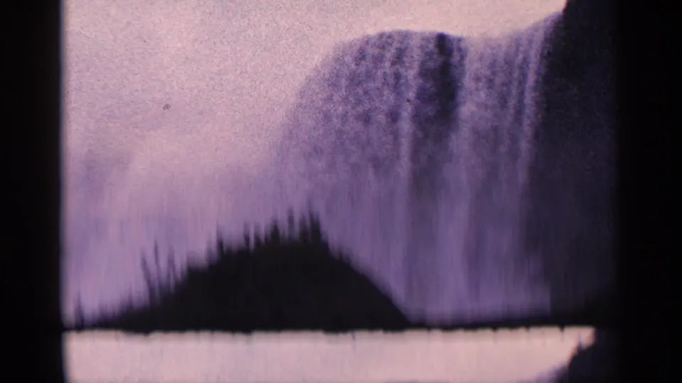1964 Niagara Falls home-movie frame with the waterfall partly obscured by a dark shoreline or treetop silhouette in the foreground.