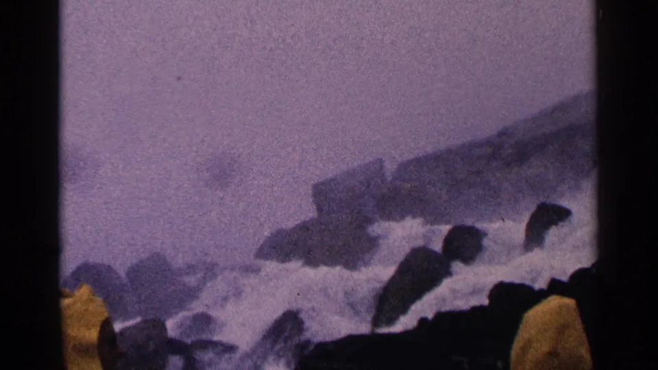 1964 Niagara Falls home-movie frame showing mist, rocks, water, and distant dark human-like silhouettes near the foreground edge.