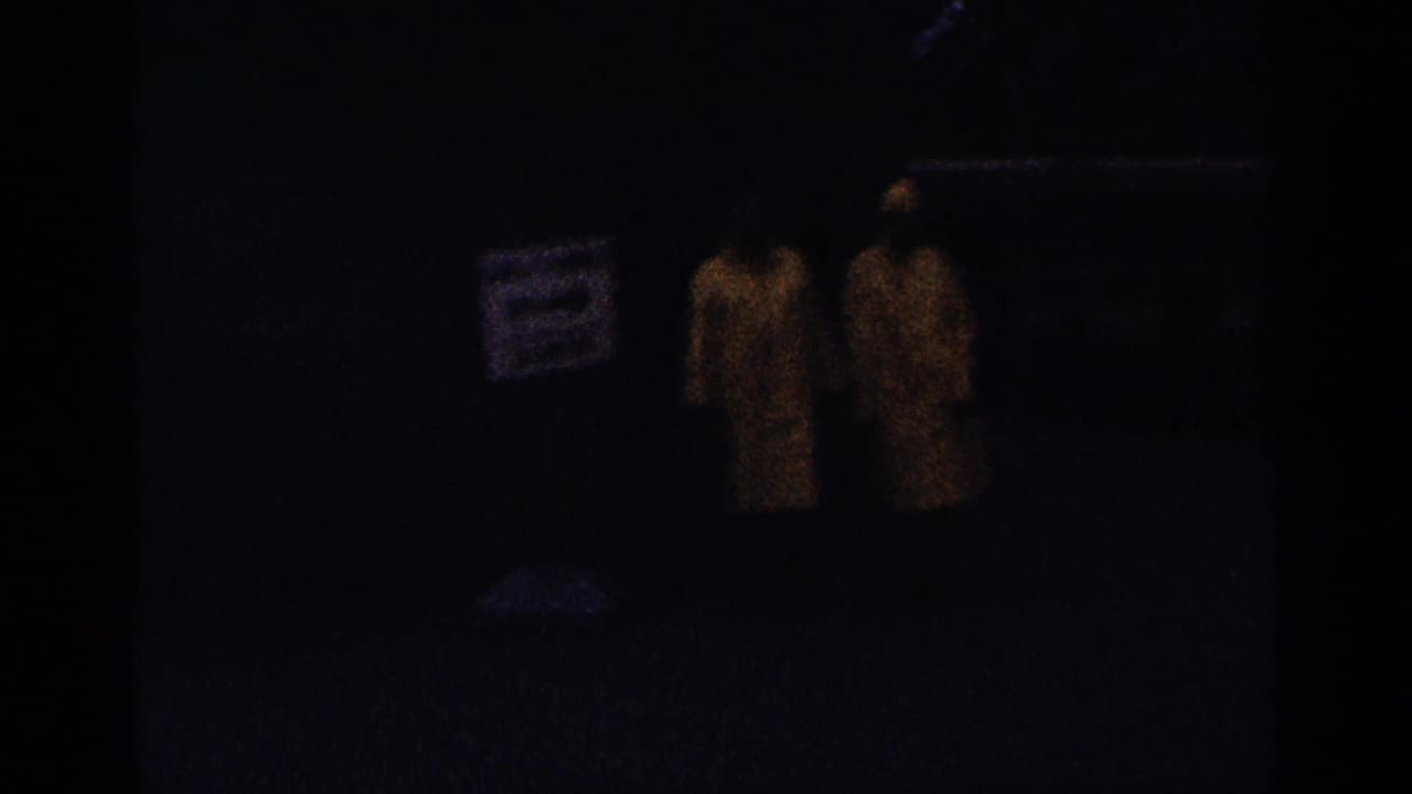 Two figures in yellow raincoats appear in a dark, grainy frame near a sign and walkway.