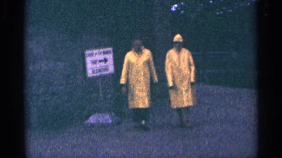 1964 Niagara Falls frame showing two adults in yellow raincoats walking along a wet path near a sign and trees.