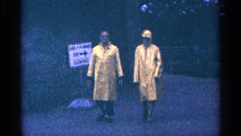 1964 Niagara Falls home-movie frame with two raincoated adults approaching on a misty walkway near a posted sign.