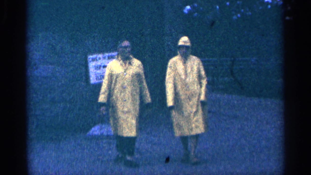 Two adults in yellow raincoats walk toward the camera in a grainy low-light scene.