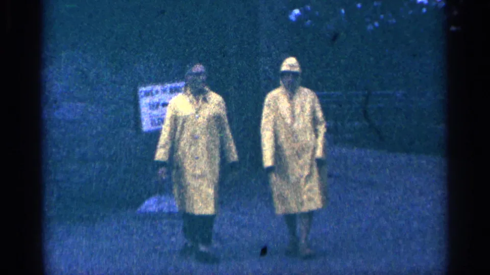 Two adults in yellow raincoats walk toward the camera in 1964 Niagara Falls home-movie footage, with a blurred sign behind them.