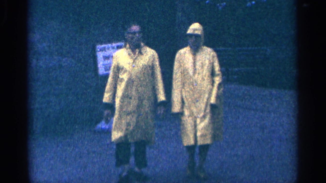 Two raincoated adults stand closer to the camera with a blurred sign in the background.