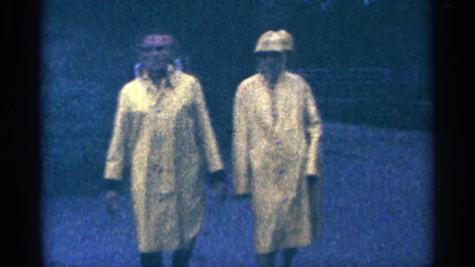 The raincoated pair fills more of the frame in a grainy 1964 Niagara Falls home-movie shot, with faces partly visible.
