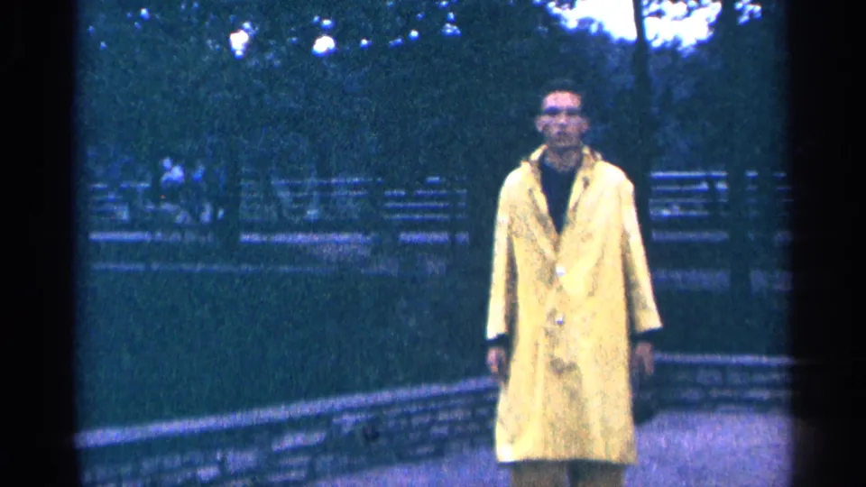 A man in a yellow raincoat faces the camera on a park path, with benches, trees, and a low wall behind him.