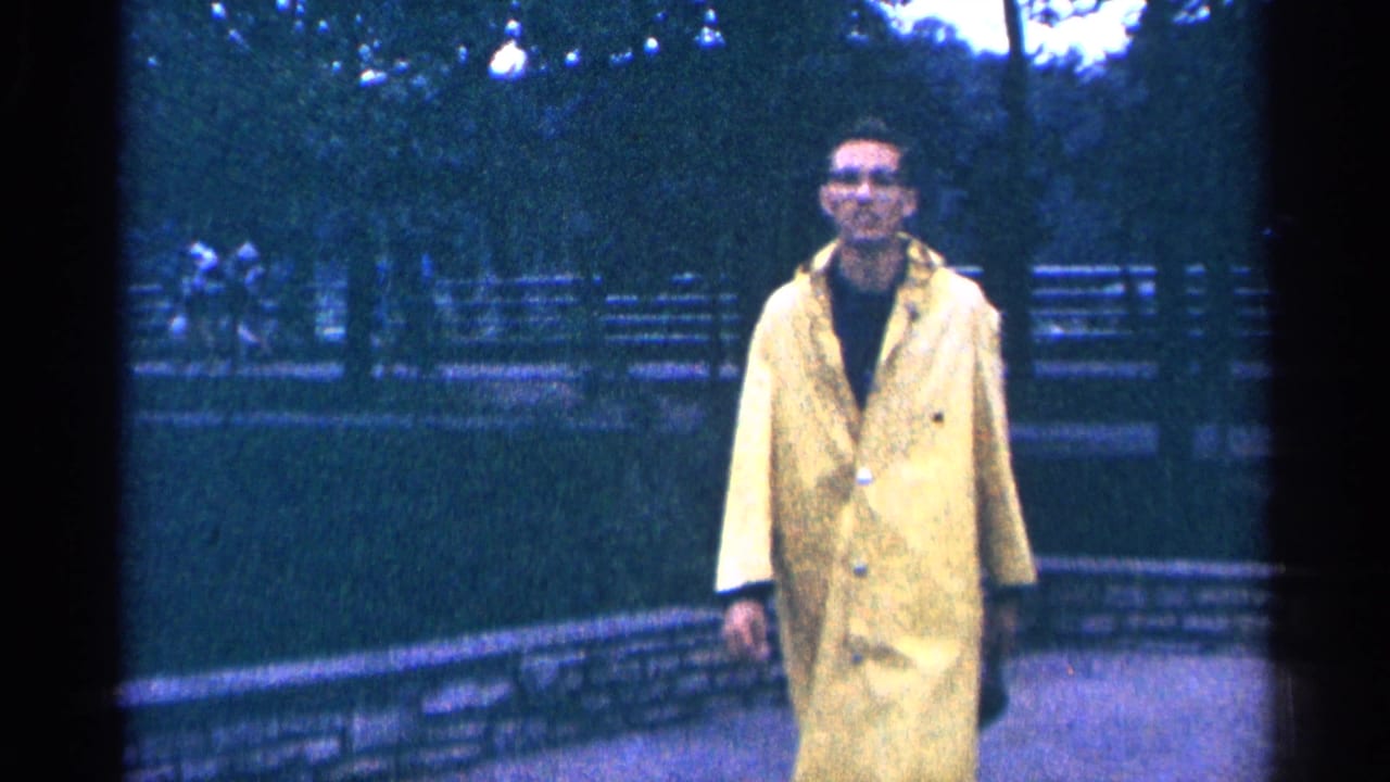 Man in a yellow raincoat walks along a park path with benches and trees behind him.