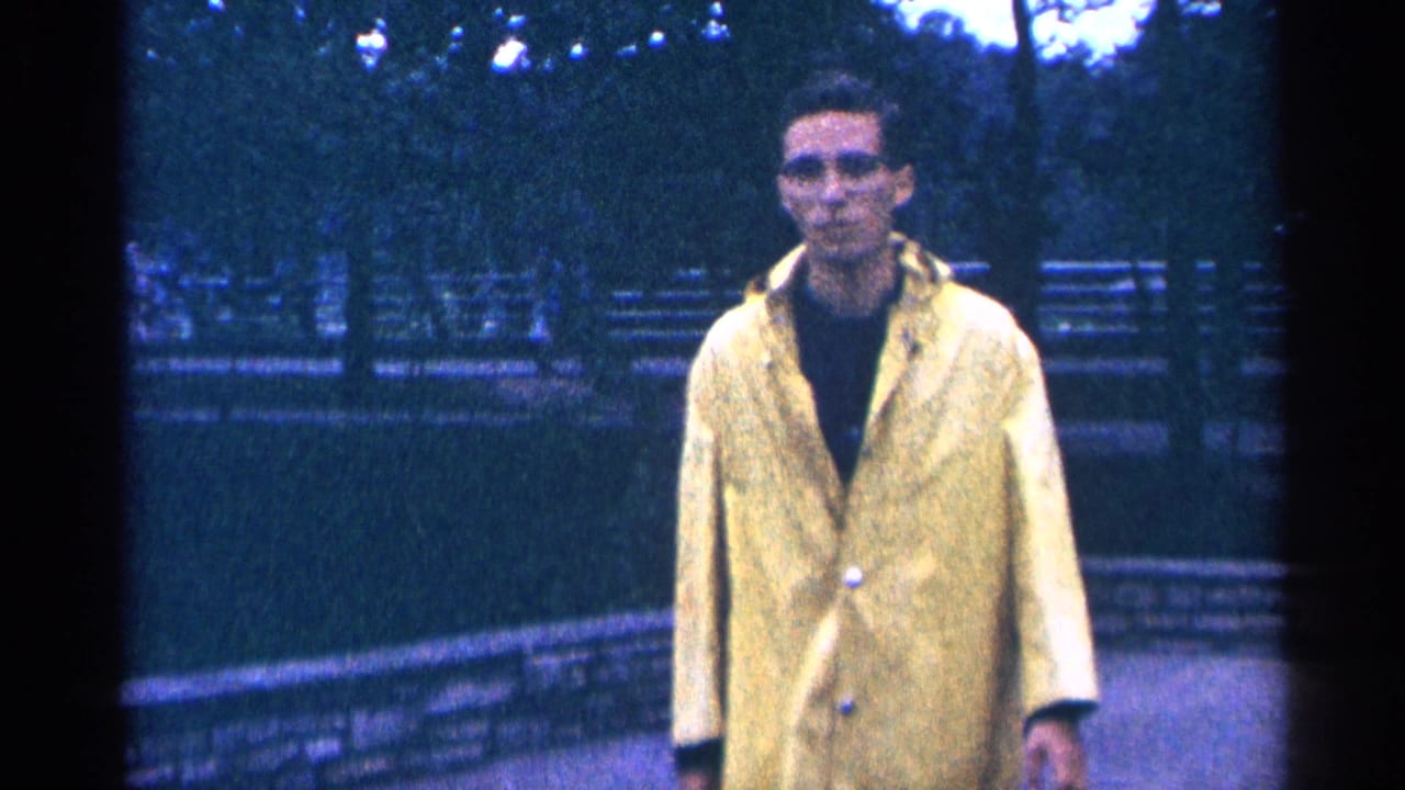 Man in a yellow raincoat stands closer to the camera on a tree-lined path.