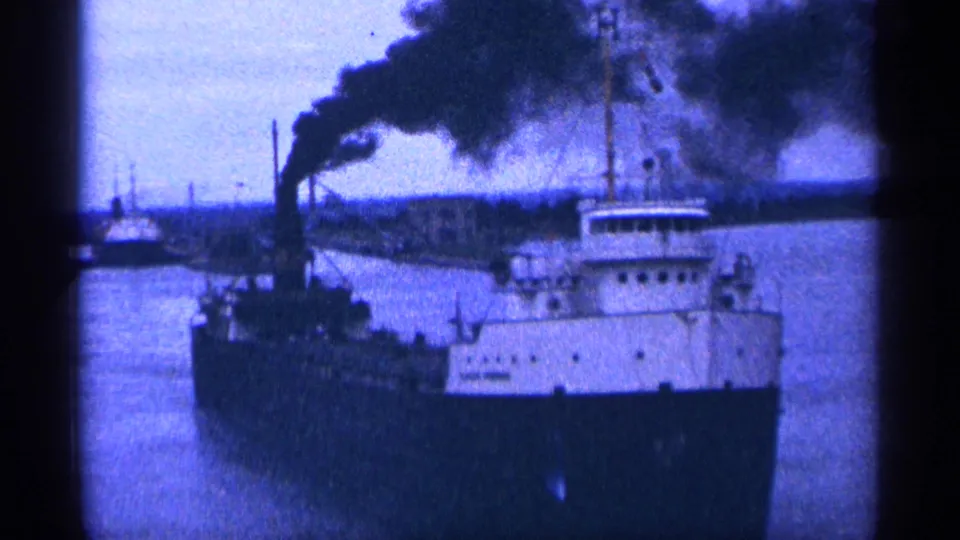 Side view of a freighter with a broad plume of black smoke over the water, filmed with blue-shifted 8mm texture.