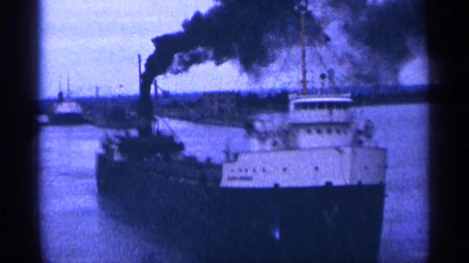 1964 home-movie frame of a large vessel moving along a waterway, with a white upper deck and dark smokestack smoke.