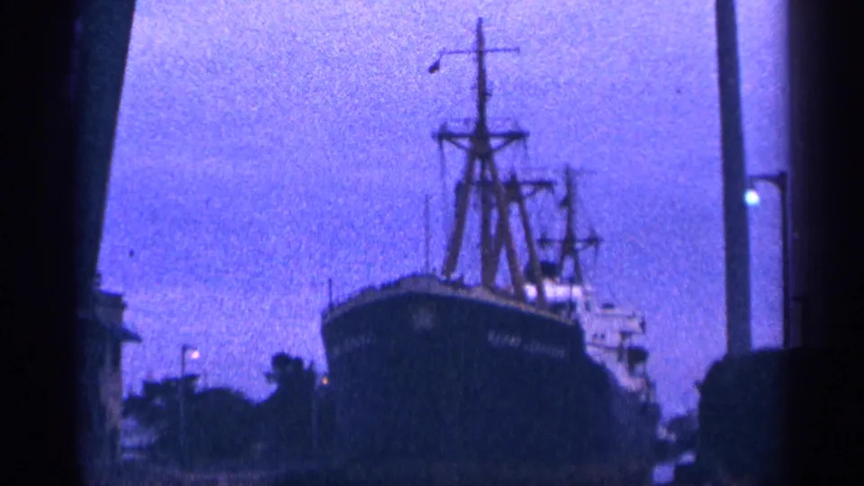 A dockside or shoreline view shows the dark bow of a large ship with tall masts and rigging against a blue-tinted sky.
