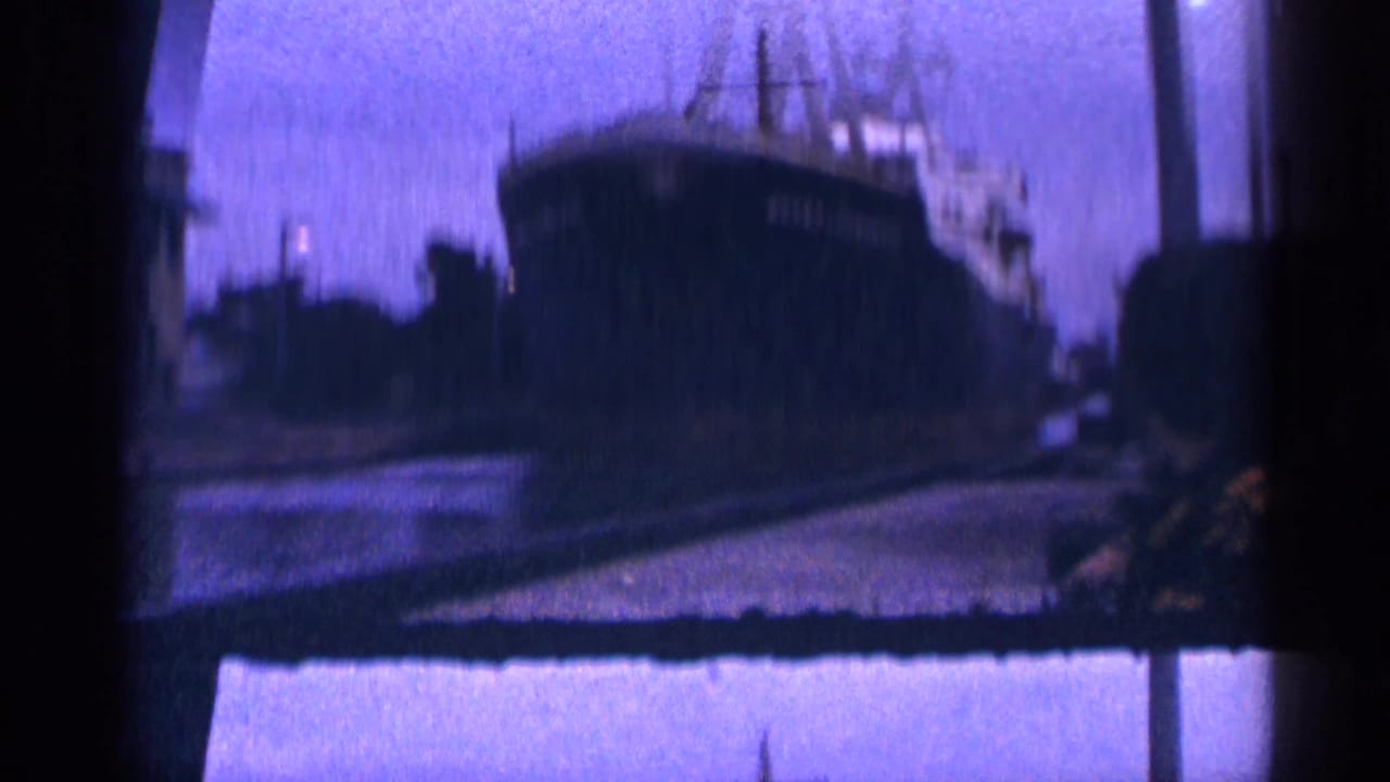 1964 8mm frame showing a large docked ship in soft focus against a blue-purple dusk sky, with dark frame edges and faint dock lighting.