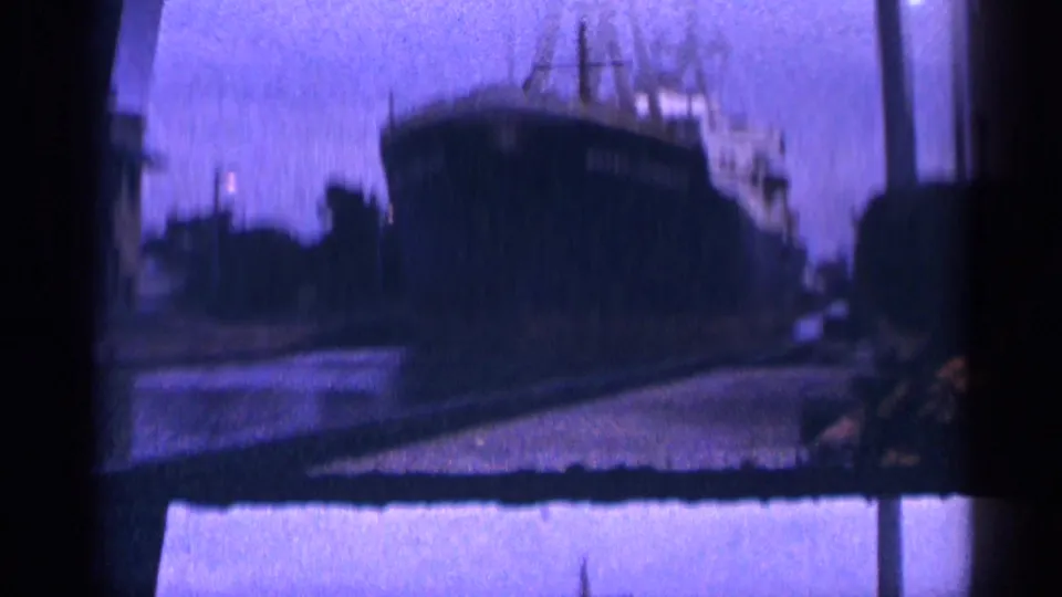 1964 8mm frame showing a large docked ship in soft focus against a blue-purple dusk sky, with dark frame edges and faint dock lighting.