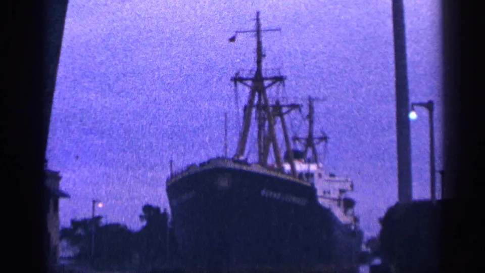 1964 8mm frame of a docked ship with tall mast structures and a dark waterfront foreground, filmed in grainy twilight color.