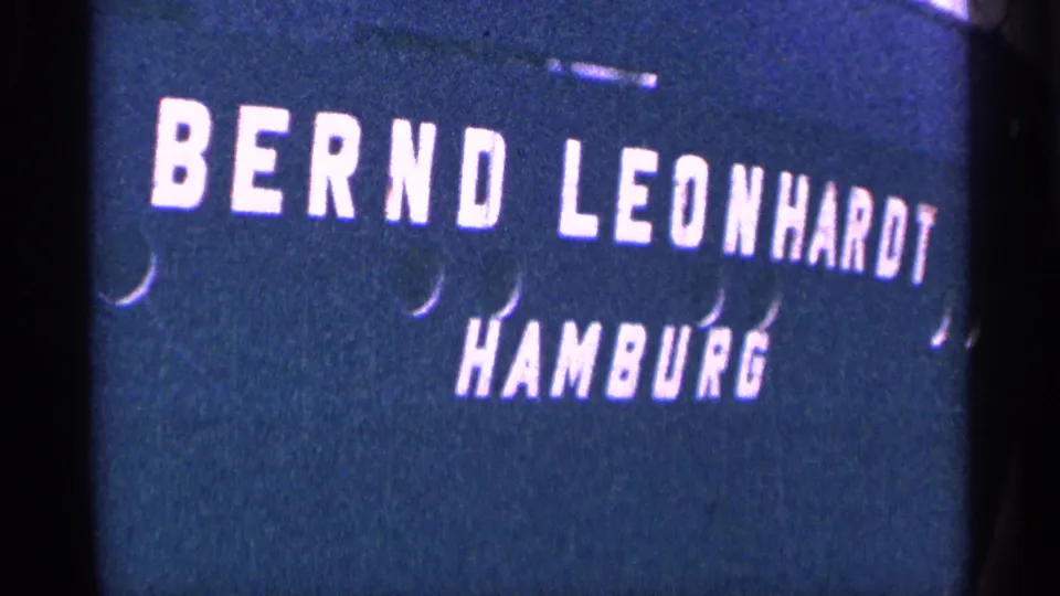 Later 8mm frame holding on the Bernd Leonhardt Hamburg title card, with soft focus, blue tint, and dark vertical film-edge shadows.