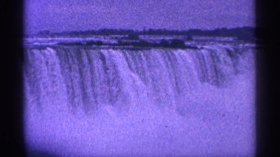 Wide 1964 8mm view of Niagara Falls with water spilling over the crest under a purple-blue sky.