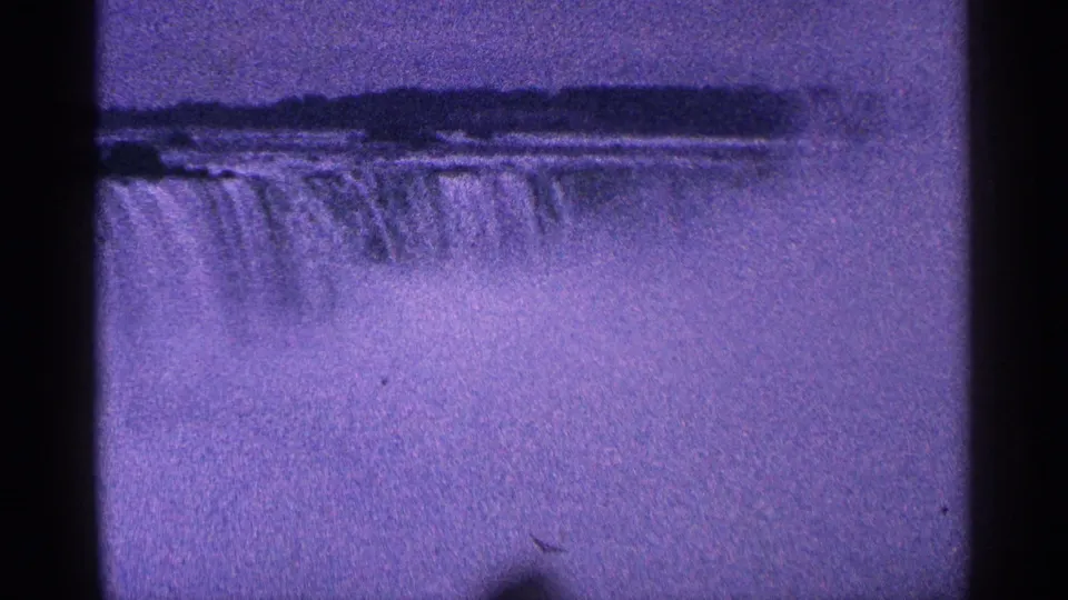 The 1964 home-movie frame shows Niagara Falls from a distance, with the waterfall edge, heavy mist, and a dark treeline under a purple cast.