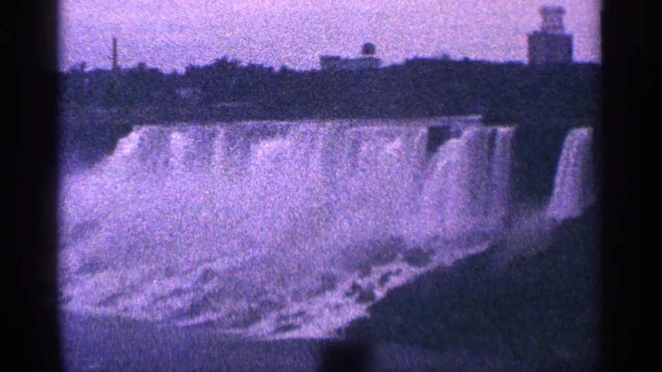 Clearer 1964 8mm view of Niagara Falls, Canada, showing the cascade, mist, shoreline, distant buildings, and a tower-like structure.