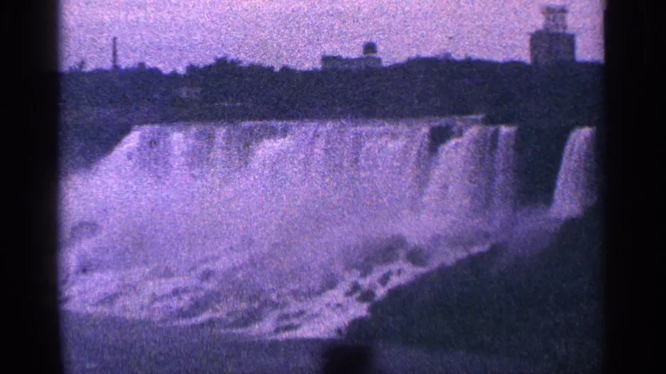 1964 8mm view of Niagara Falls in Canada, showing the waterfall edge, spray, and distant structures through a purple-tinted film scan.