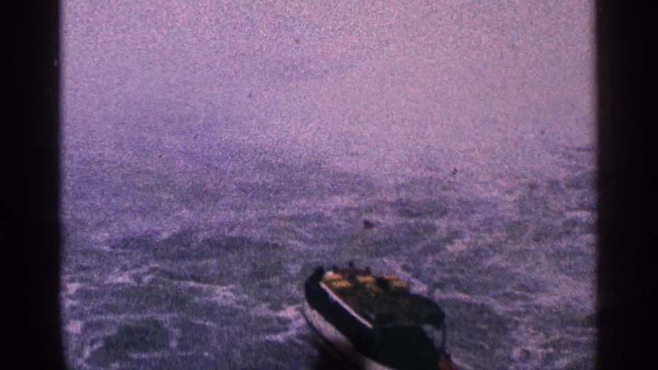 A small dark boat sits low in the 8mm frame, surrounded by grey-blue chop and soft mist, with passengers not individually identifiable.