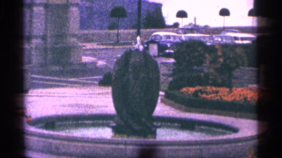 1964 Niagara Falls, Canada home-movie frame of a landscaped fountain, flower bed, walkway, and parked mid-century cars.