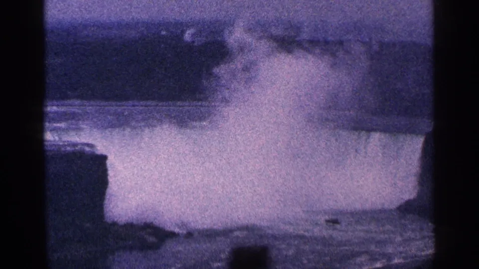 1964 8mm home-movie frame of Niagara Falls, Canada, showing mist rising from cascading water beside dark rock forms.