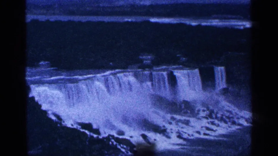 1964 home-movie frame of Niagara Falls from a distant overlook, showing cascading water, mist, rocks, and dark gorge areas.