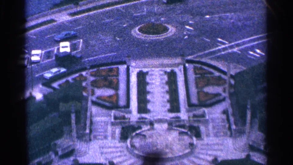 Blue-tinted 8mm frame of a Niagara Falls street layout with cars, a circular island, formal beds, stairways, and a plaza fountain.