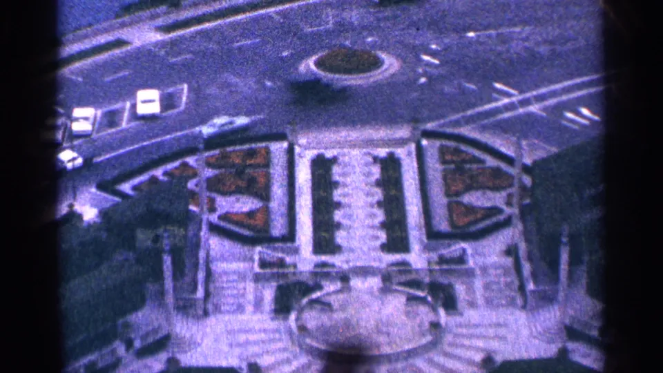 1964 Niagara Falls home-movie frame showing cars on a curved roadway around a traffic island, with formal landscaping and a fountain below.